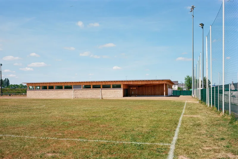 Low pavilion with timber cladding and clerestory windows beside a grass sports field under a clear sky