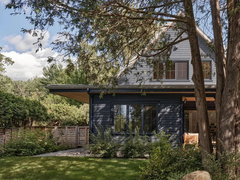 Side view of the two-storey volume with dark cladding below and pale siding above under mature pines