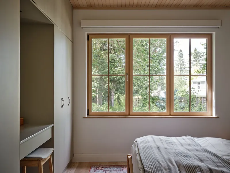 Bedroom with timber-framed casement windows overlooking summer foliage and neighboring houses
