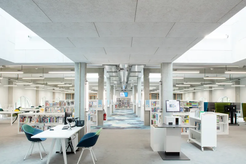 Library interior with concrete columns, exposed ductwork and white bookshelves under a coffered concrete ceiling