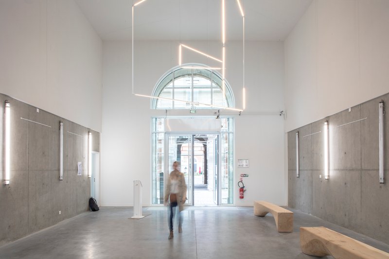 Entry hall with concrete wall panels, linear pendant lighting and a figure walking toward the arched doorway