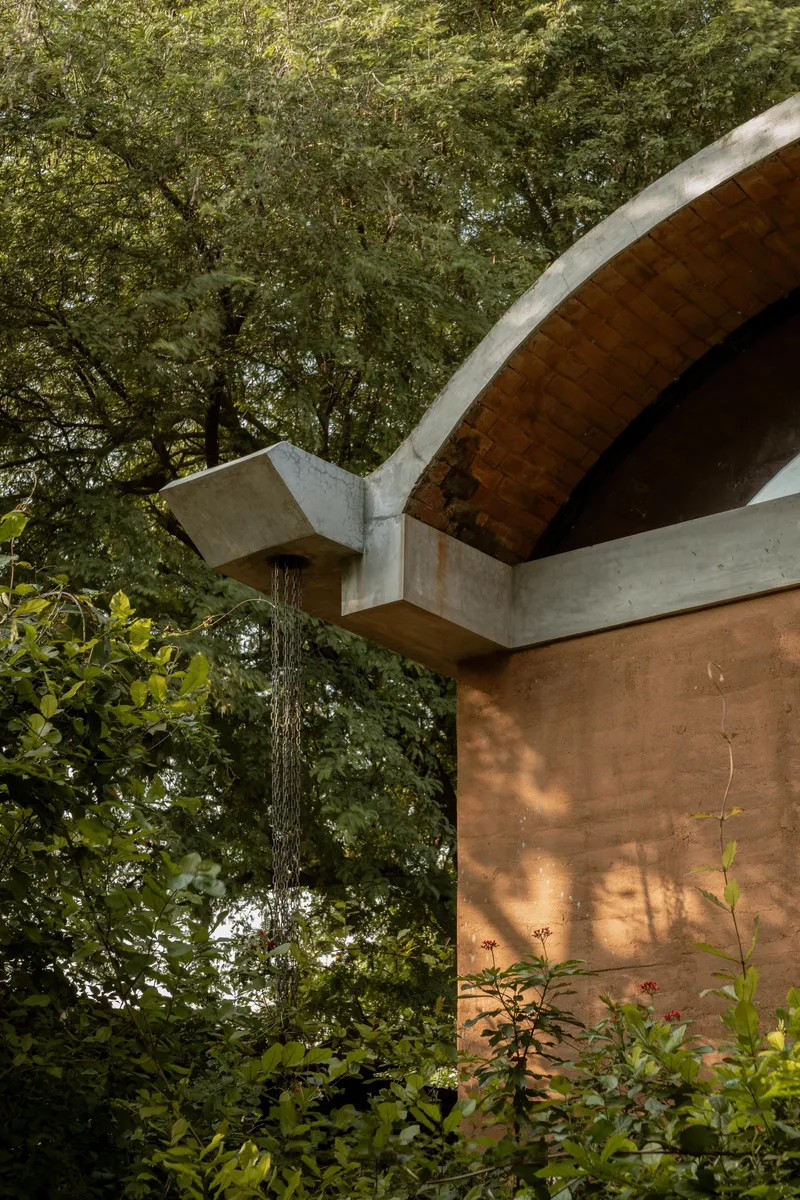 Concrete barrel vault roof soffit meeting a rammed earth wall beneath dappled tree canopy shade