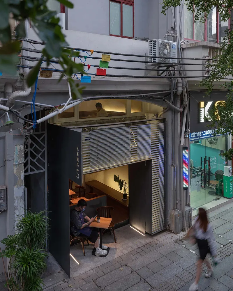 Two-story shopfront with horizontal louvers and outdoor seating as pedestrians pass in early evening