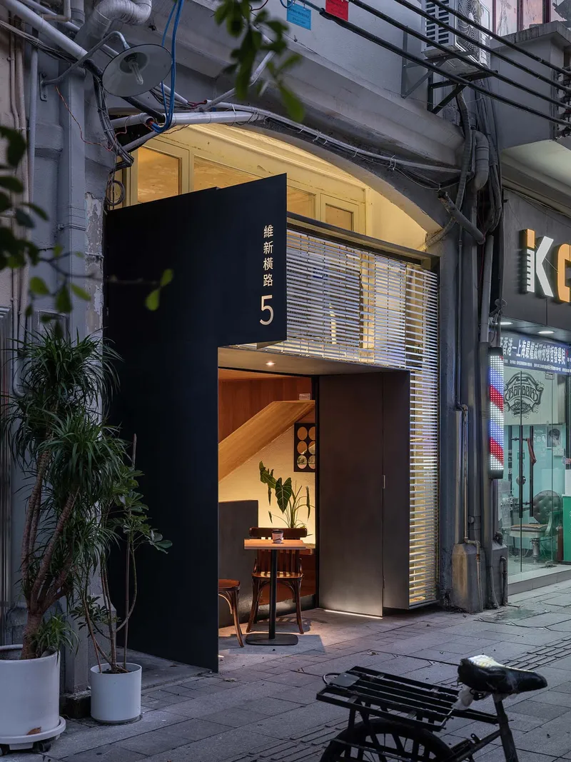 Narrow alley entrance with black facade panel and horizontal louvers at twilight beside potted plants