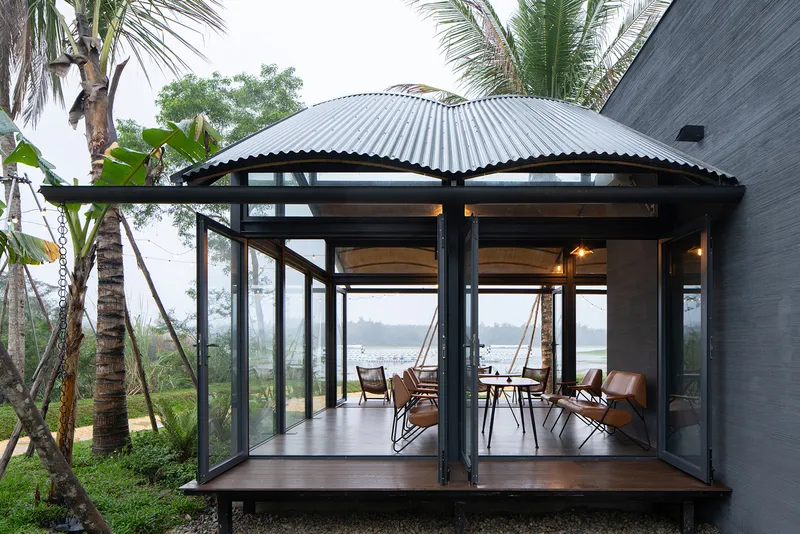 Glazed pavilion with corrugated metal roof and black steel frame nestled among palm trees overlooking the water