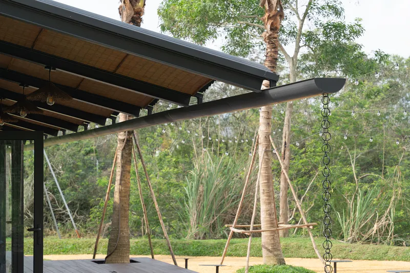 Detail of rope swings hanging from the timber-lined roof overhang with staked palm trees in the landscape beyond