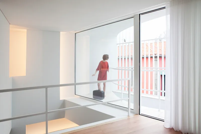 Double-height space with child standing on balcony threshold overlooking street through glass door