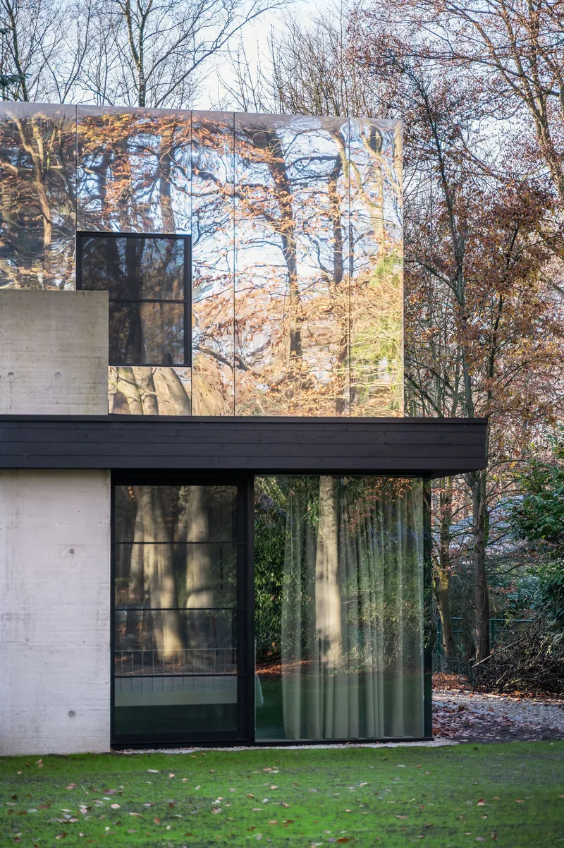Corner detail of mirrored upper volume reflecting bare branches above dark-framed sliding glass doors