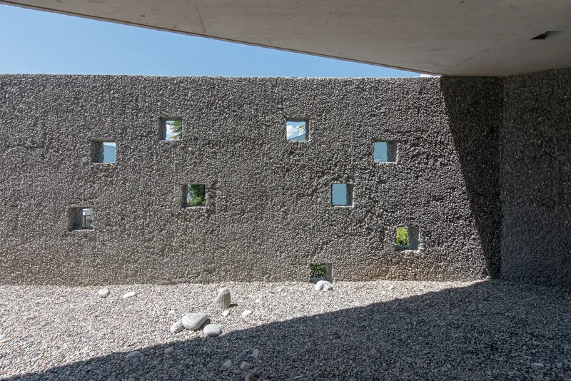Exterior gravel courtyard wall with scattered square window openings beneath concrete overhang