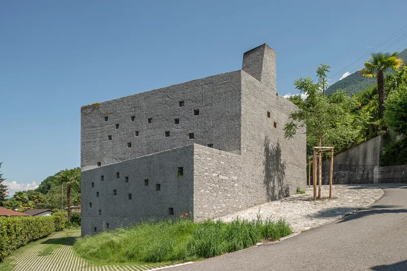 Stacked concrete volumes with punched square windows on a sloping roadside with palm trees and mountains