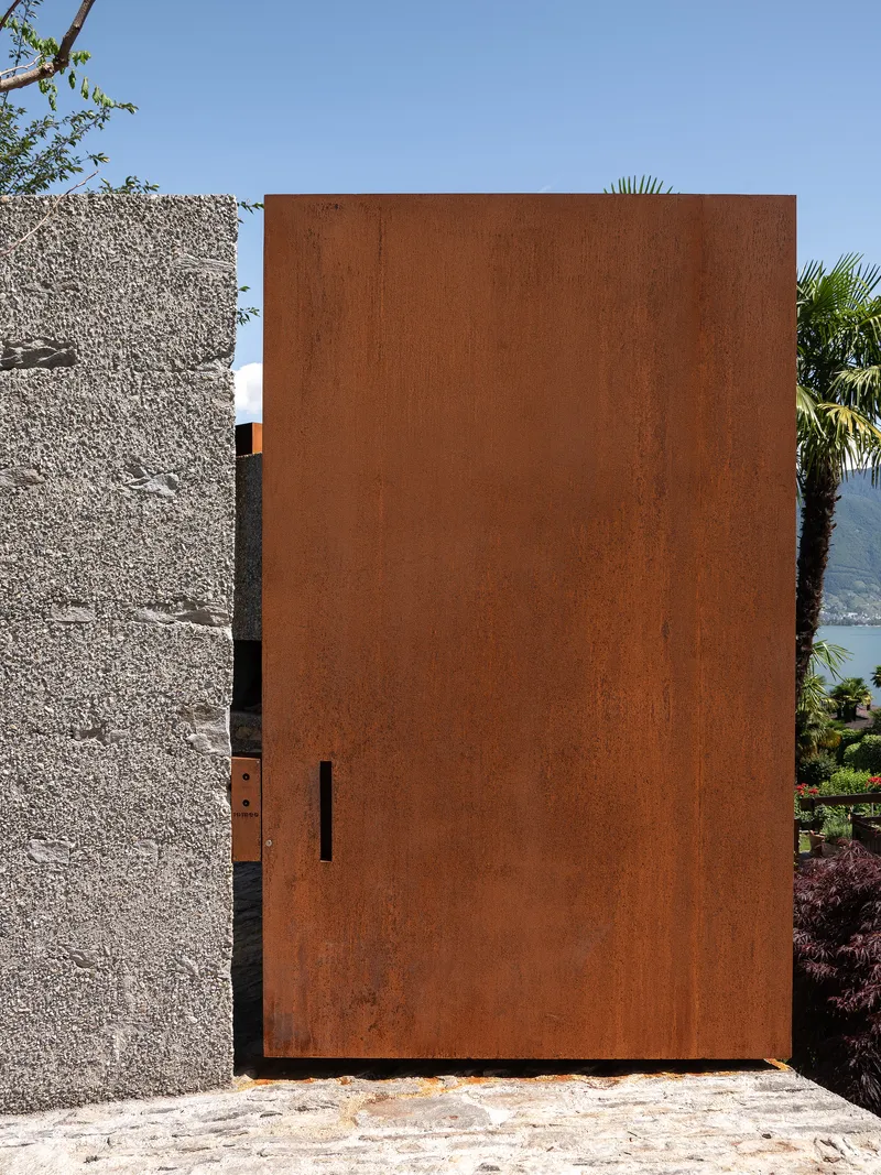 Weathered steel entry door panel with narrow vertical slot flanked by exposed aggregate concrete walls