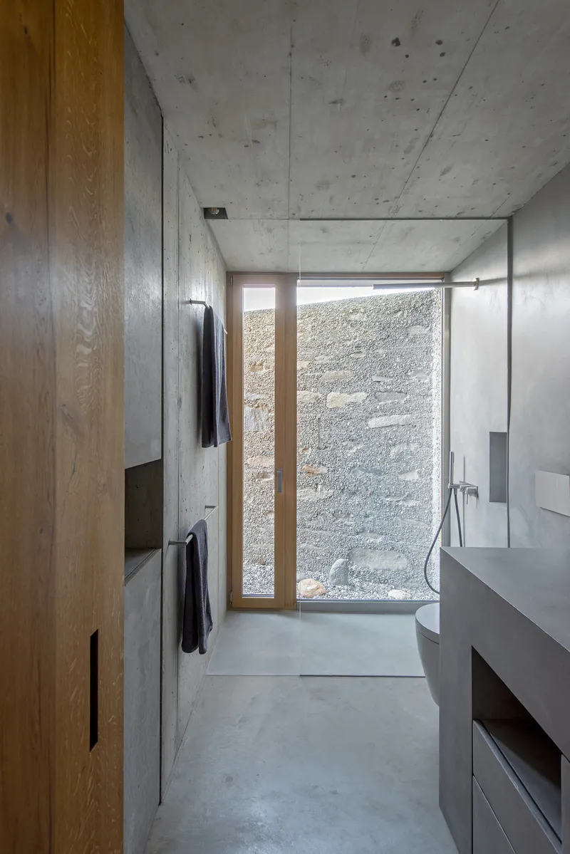 Bathroom with concrete walls, timber cabinetry, and a narrow window facing the textured courtyard wall