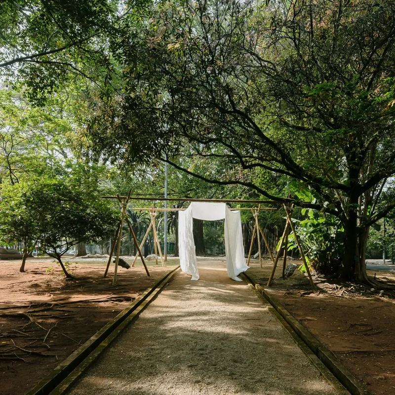 Timber frame pavilion with white fabric panels along a paved pathway beneath a canopy of trees