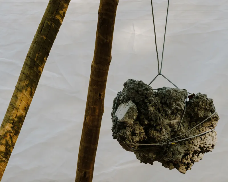 Concrete block suspended by steel cables between two bamboo poles with white fabric in the background