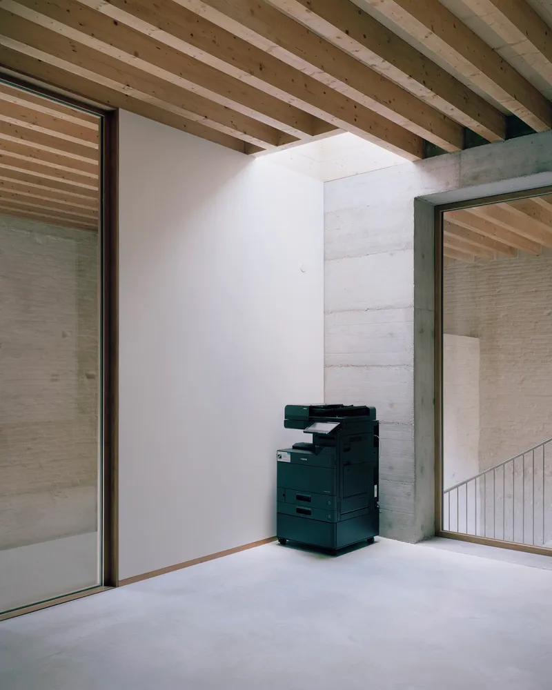 Corner nook with exposed timber joists overhead and printer stationed against white partition near glazed opening
