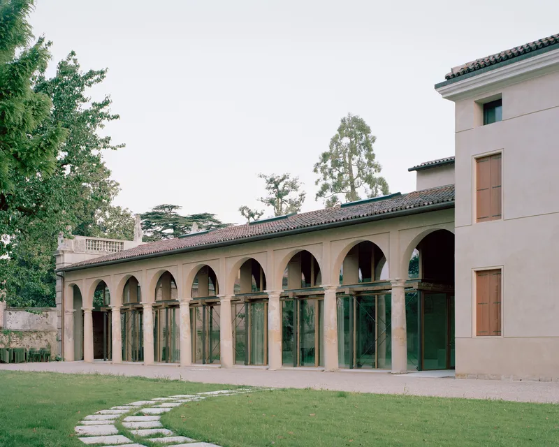 Arcaded loggia with glass infill and pink stucco volumes set behind a stone-paved lawn path