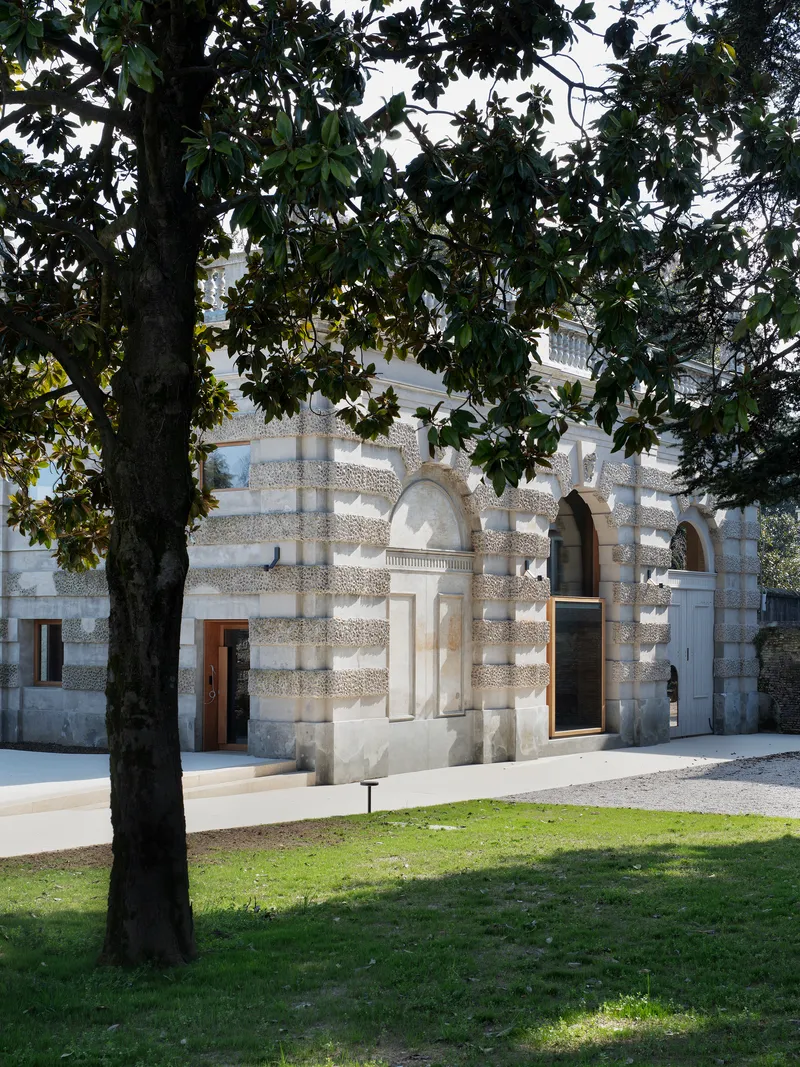 Stone facade with arched openings and balustrade viewed through overhanging tree branches in afternoon light