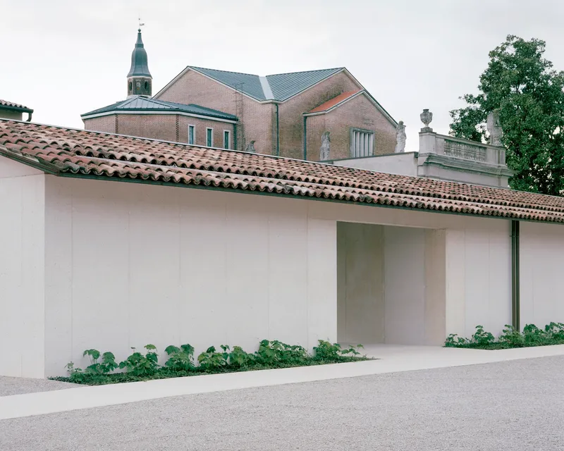White plastered perimeter wall with tiled roof and recessed entry, with church campanile visible beyond
