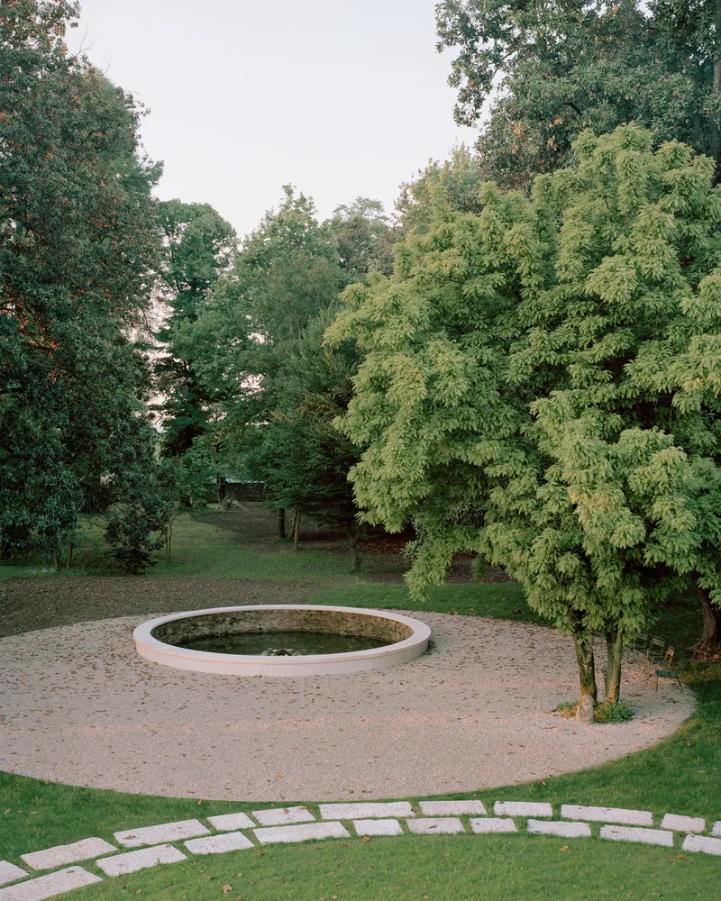 Circular stone fountain in a gravel courtyard surrounded by lawn and deciduous trees