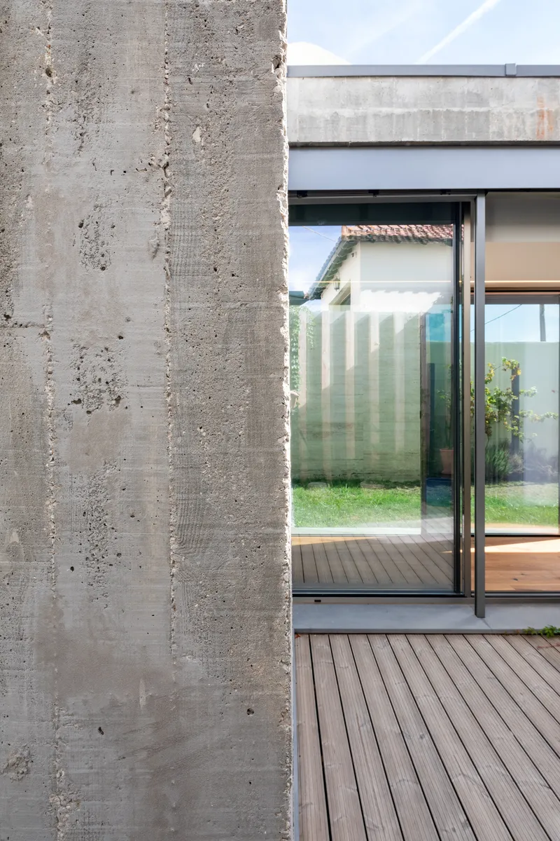 Detail of board-formed concrete wall corner meeting glass doors and timber decking with garden visible beyond