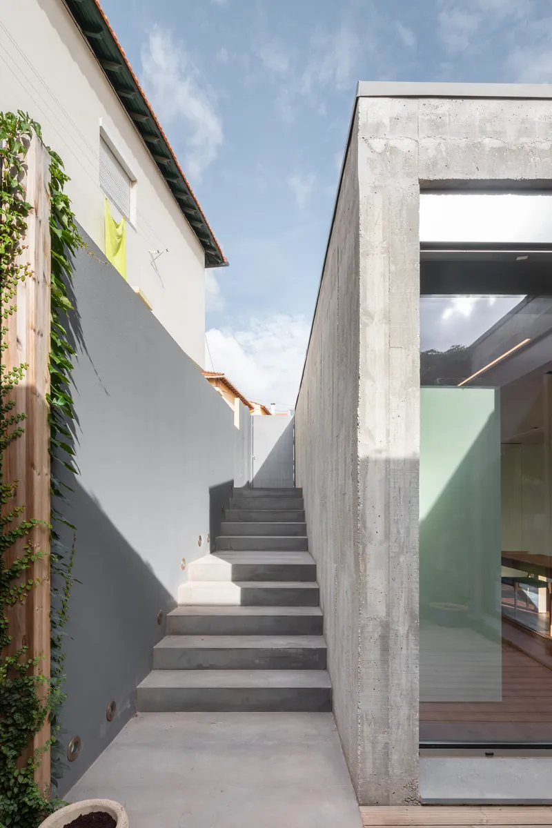 Concrete staircase between boardform concrete walls and an existing white rendered building with climbing vines