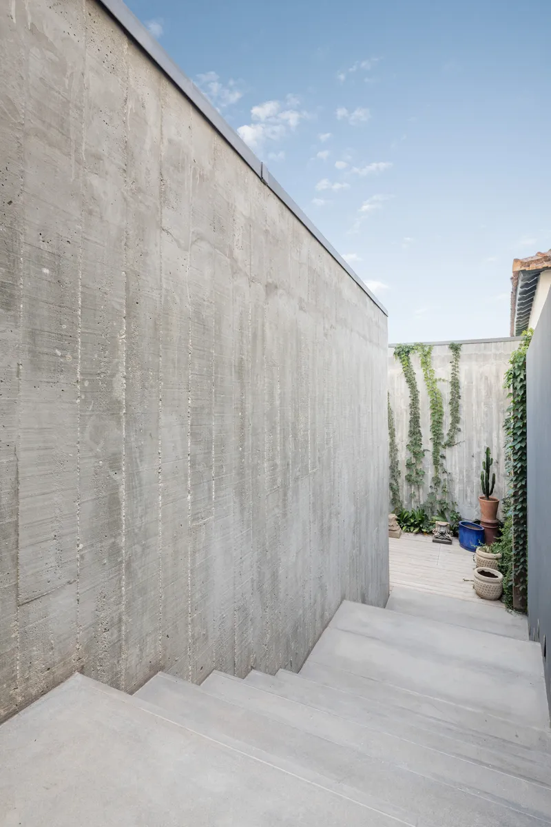 Boardform concrete wall descending with exterior stairs into a planted courtyard under blue sky