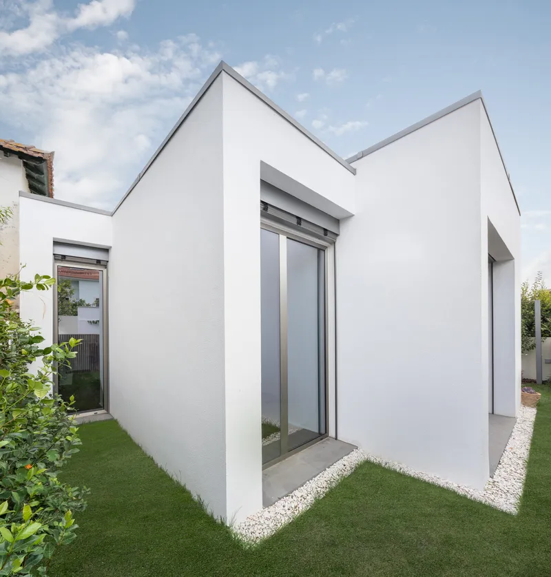 White rendered gabled volumes with sliding glass doors opening onto a lawn under scattered clouds