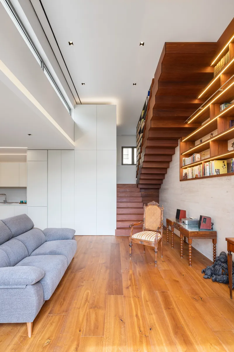 Open living space with floating stair linking timber shelving wall and white kitchen under clerestory windows