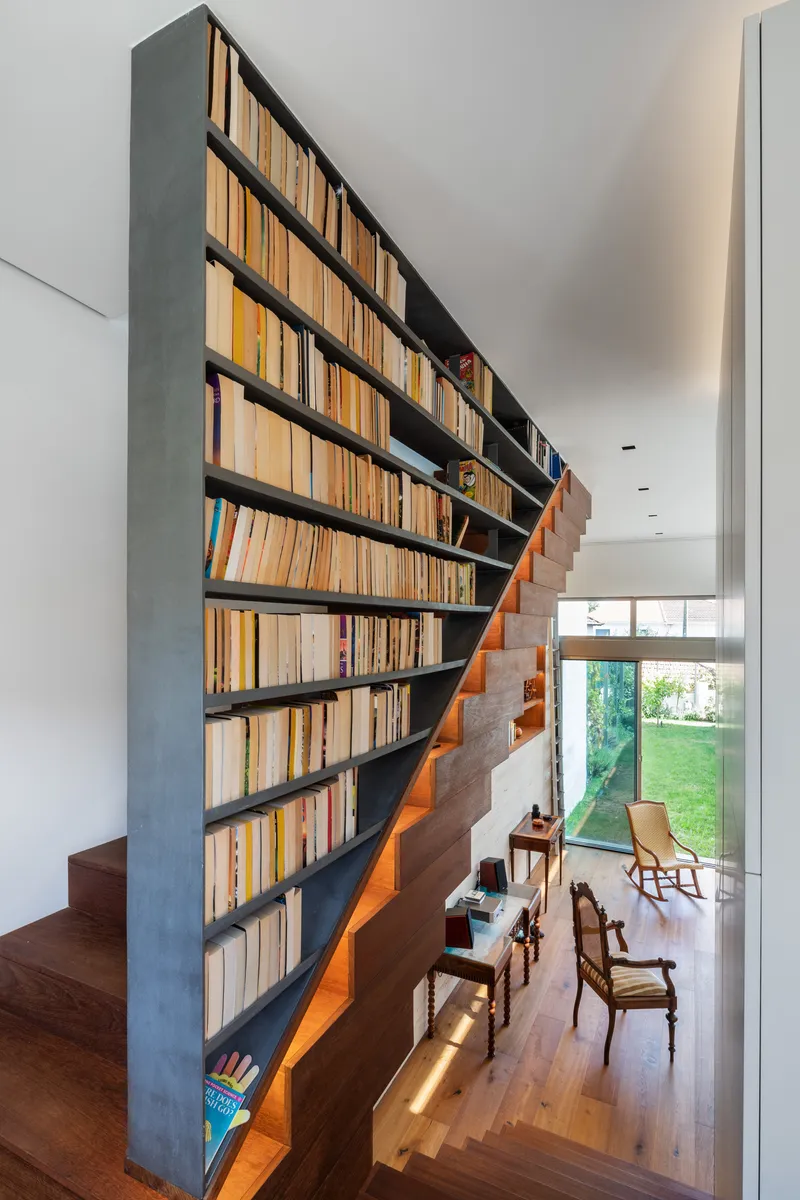 Upper-level view down metal and timber bookshelf wall beside cantilevered staircase with garden beyond