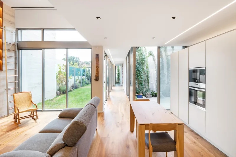 Long interior corridor connecting living and kitchen zones with planted courtyards visible through floor-to-ceiling glass