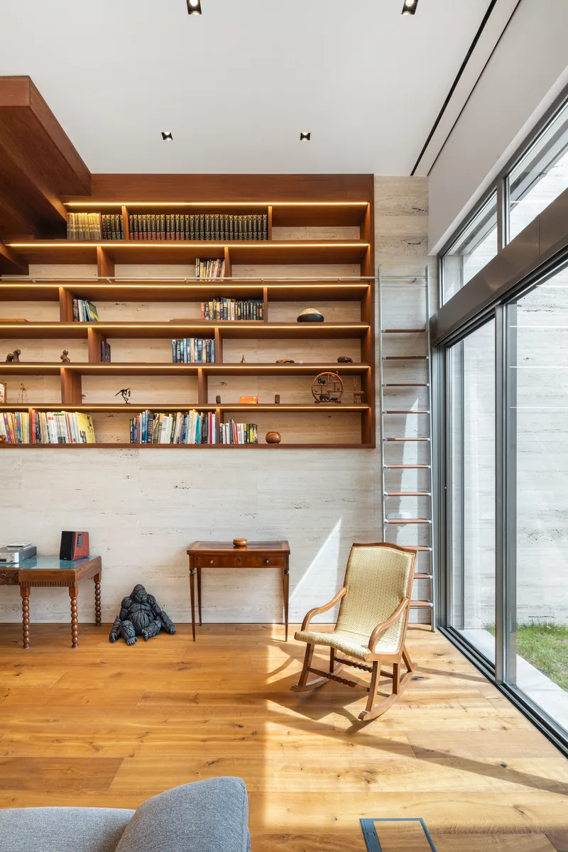Timber bookshelves with library ladder and wide-plank oak flooring in afternoon sunlight beside tall glazed wall