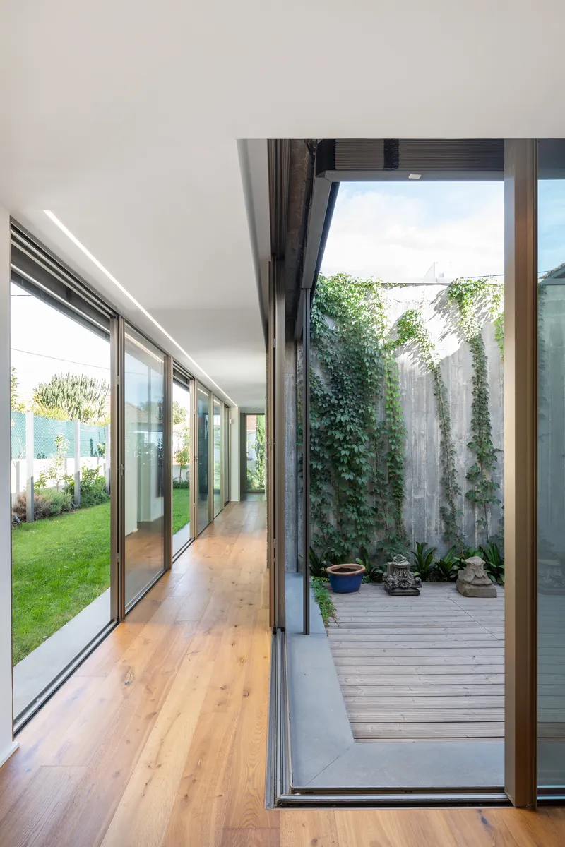 Interior corridor with timber flooring and floor-to-ceiling glazing opening onto a timber-decked courtyard with climbing vines