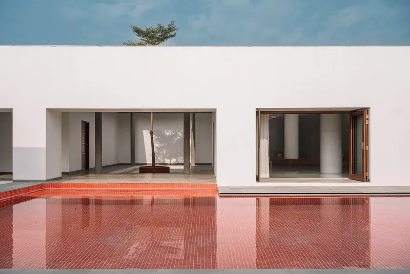 Red mosaic tile pool reflecting white stucco volumes with timber-framed openings beneath a tree canopy
