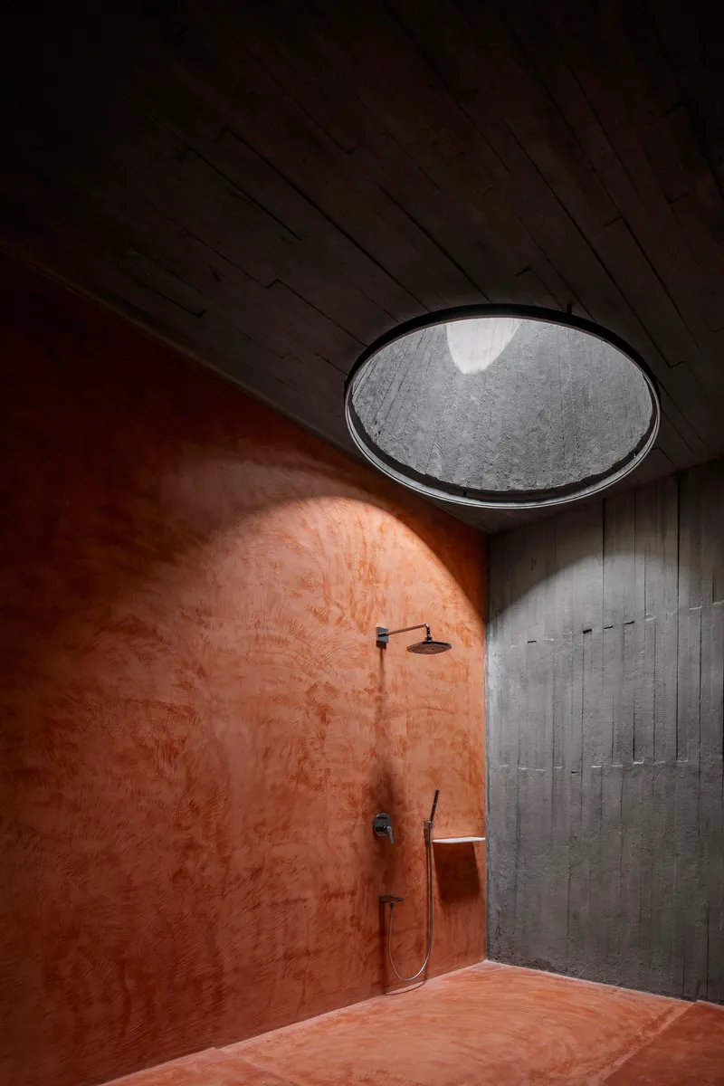 Shower alcove with circular skylight casting daylight onto terracotta plaster walls and concrete ceiling