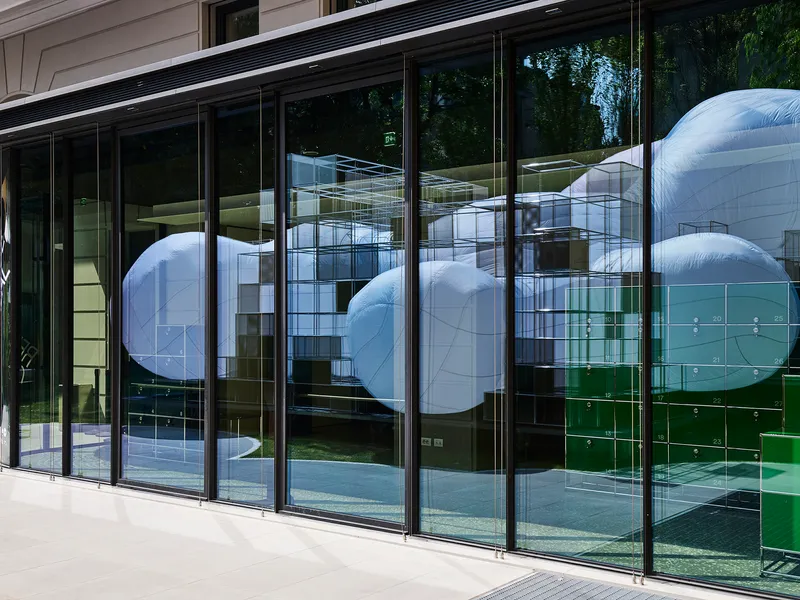 Glass facade reflecting the metal grid structure and inflatable forms with trees visible beyond
