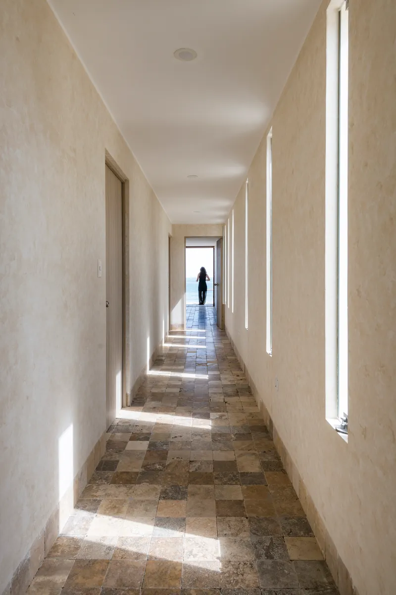 Narrow corridor with stone tile floor and vertical light slots as a figure exits into brightness