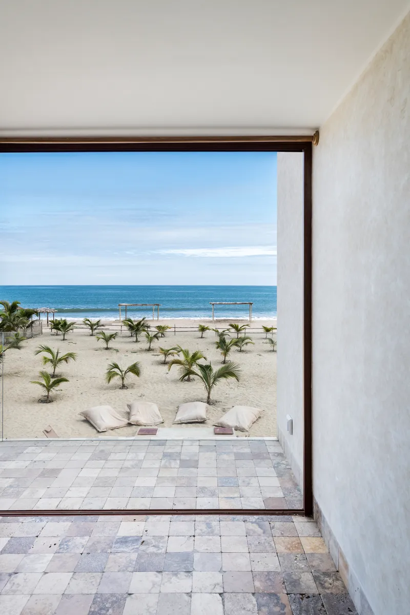 Floor-to-ceiling opening framing a beachfront with young palms, cushions, and ocean beyond
