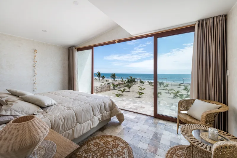 Bedroom with floor-to-ceiling glazing framing ocean views and young palm trees in the sand