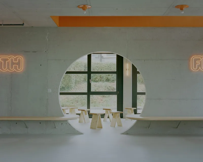 Circular portal framing a window mullion and plywood stools beneath orange neon lettering on concrete