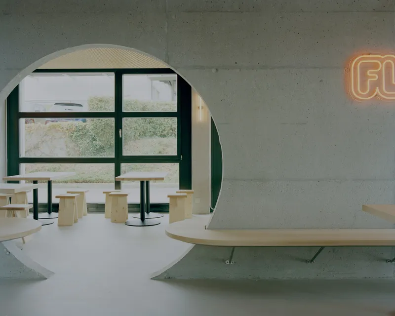 Circular arched opening framing light wood seating and dark-framed windows under a concrete ceiling