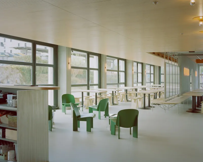 Interior with green chairs scattered among light wood furniture beneath a row of dark-framed windows