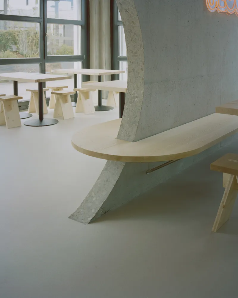 Light wood bench with angled concrete support beside plywood seating and tables near a window