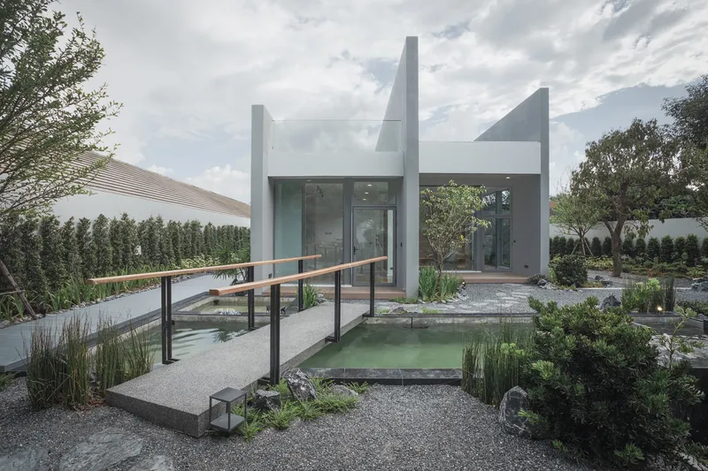 Gravel courtyard with reflecting pond and timber bridge leading to white plastered volumes under clouded skies