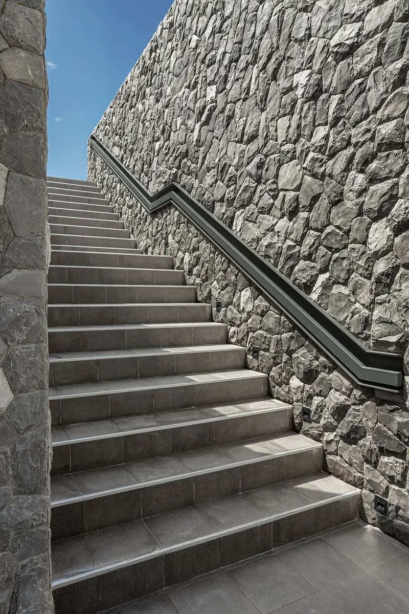 Concrete staircase with metal handrail ascending between stone-clad columns and retaining wall