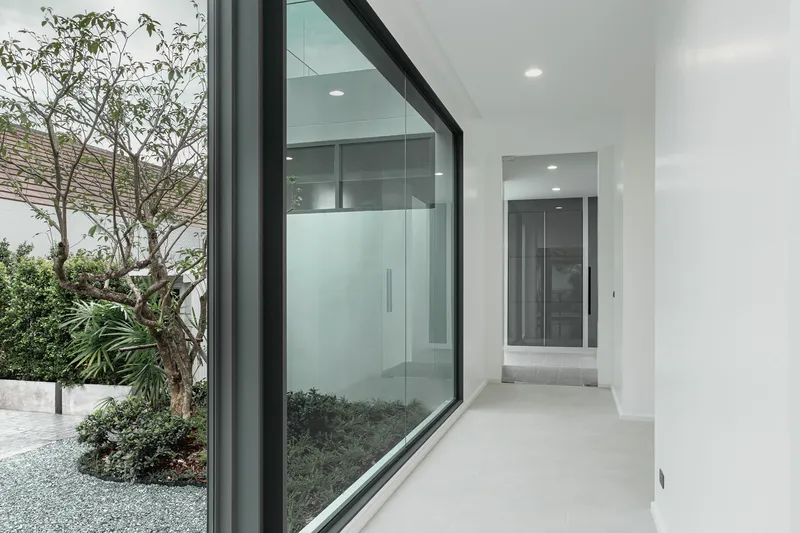White corridor with black-framed glazing opening onto a courtyard with gravel and planted trees