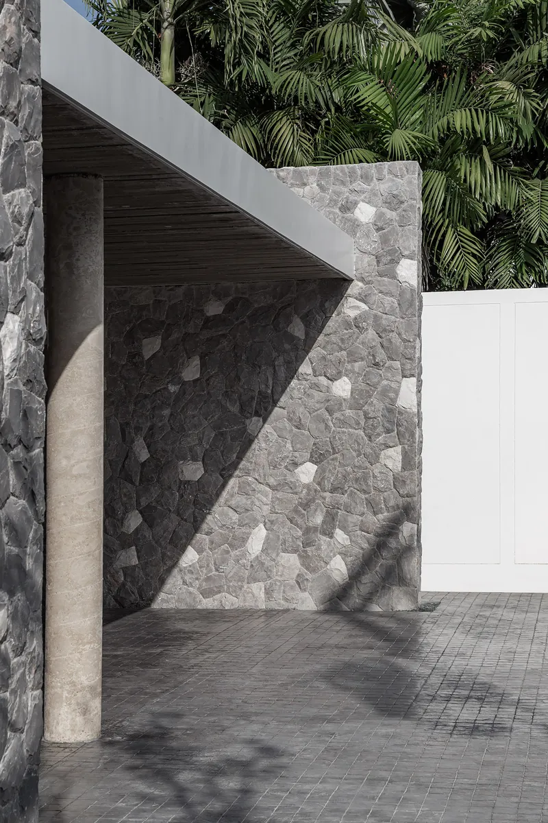 Concrete column beside a stone mosaic wall beneath a timber soffit casting angular shadows
