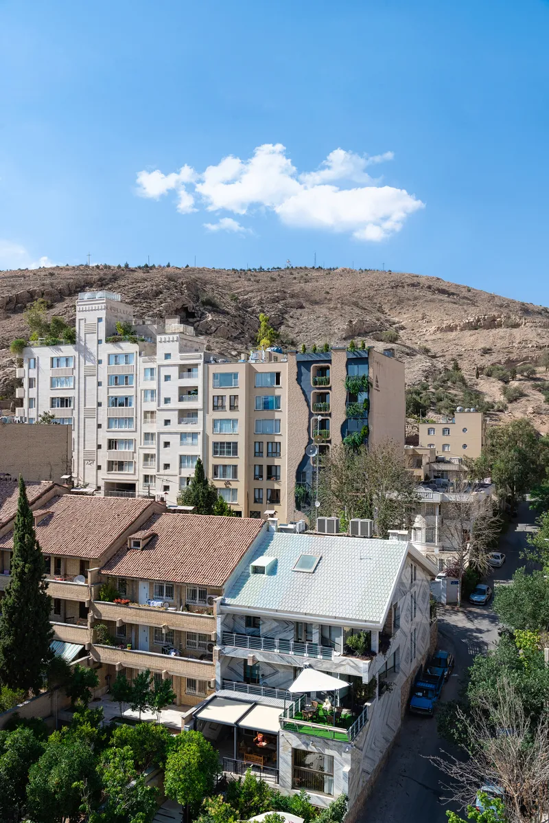 Residential tower with planted balconies rising above rooftops against a dry hillside under scattered clouds