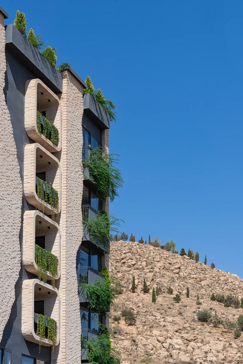 Corner view of planted balconies on a brick tower with a barren hillside beyond under clear blue sky