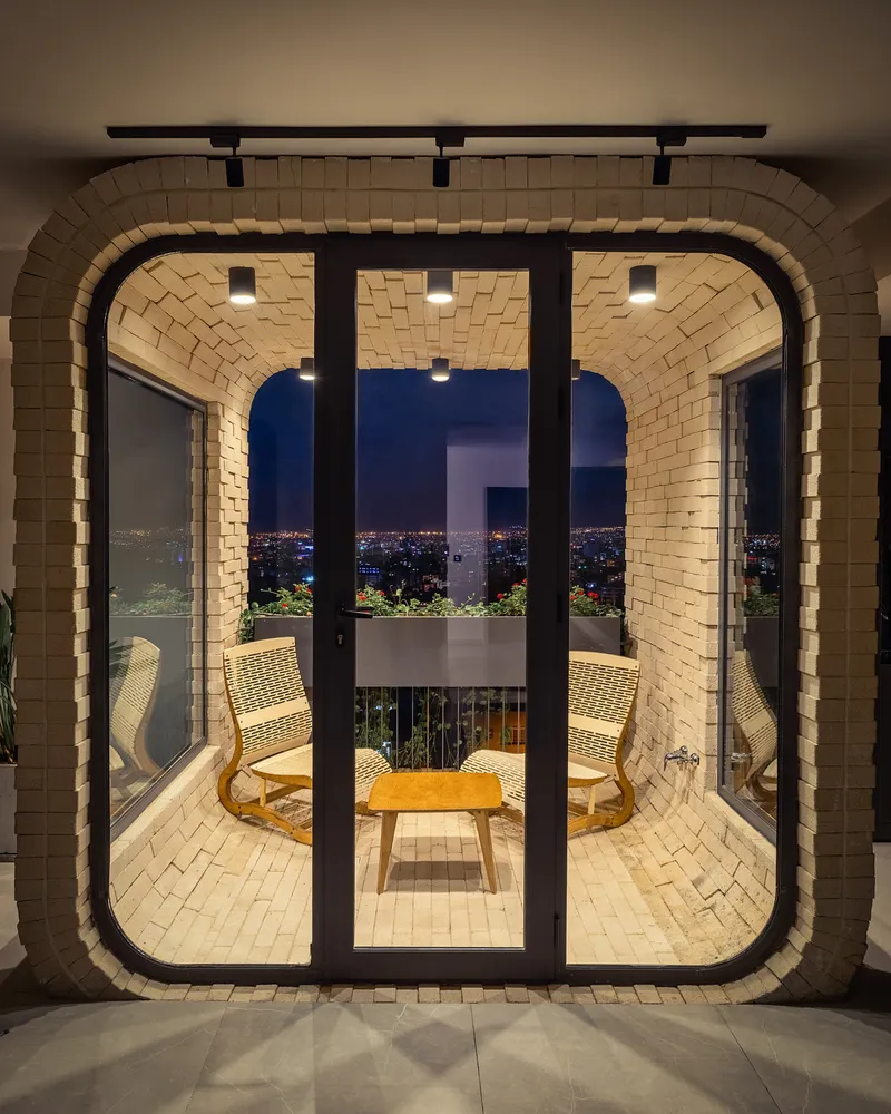 Vaulted brick pavilion with black-framed glass doors opening to a balcony overlooking city lights at dusk