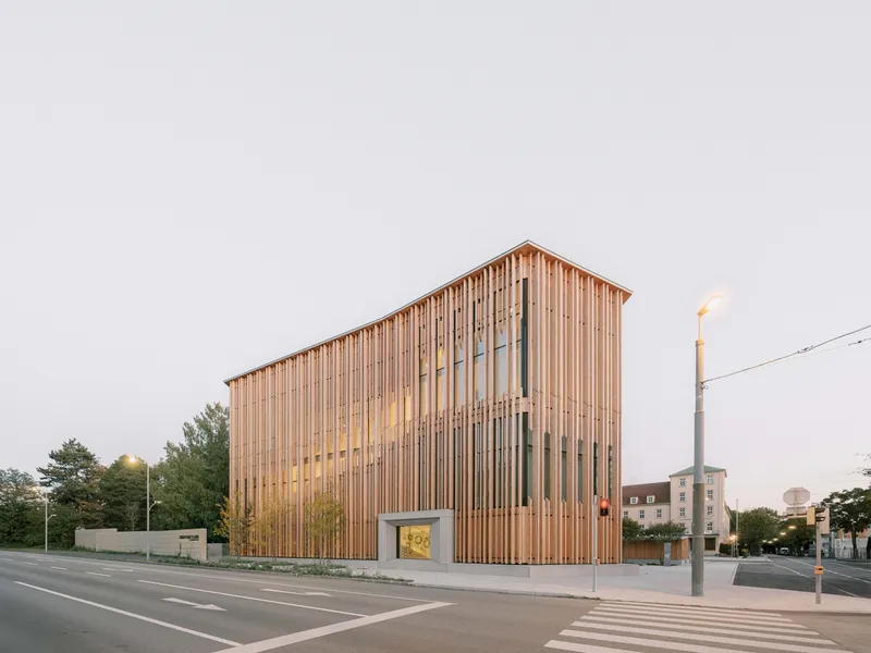 Corner elevation showing the slatted timber box facade at dusk with internal lights glowing